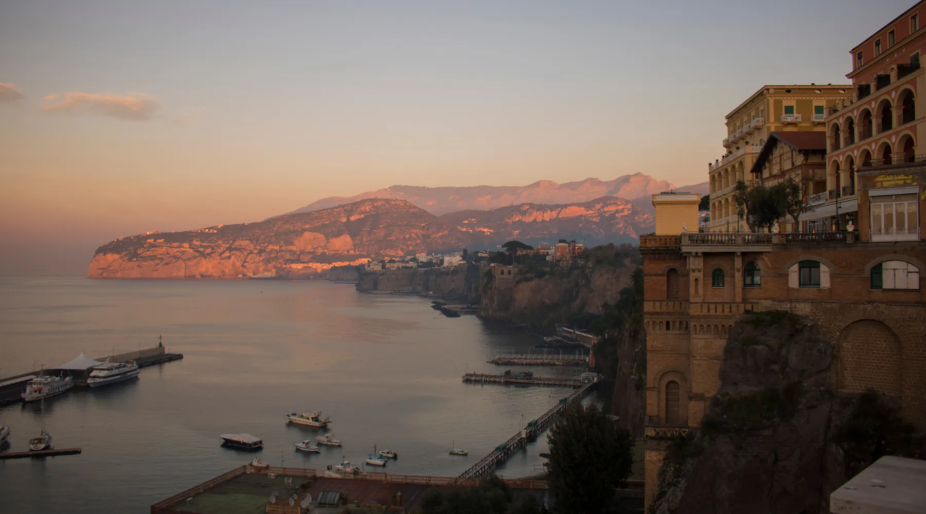 The cliffs and harbour of Sorrento at golden hour, pastel hotel façades on the right and the bay glowing pink beyond
