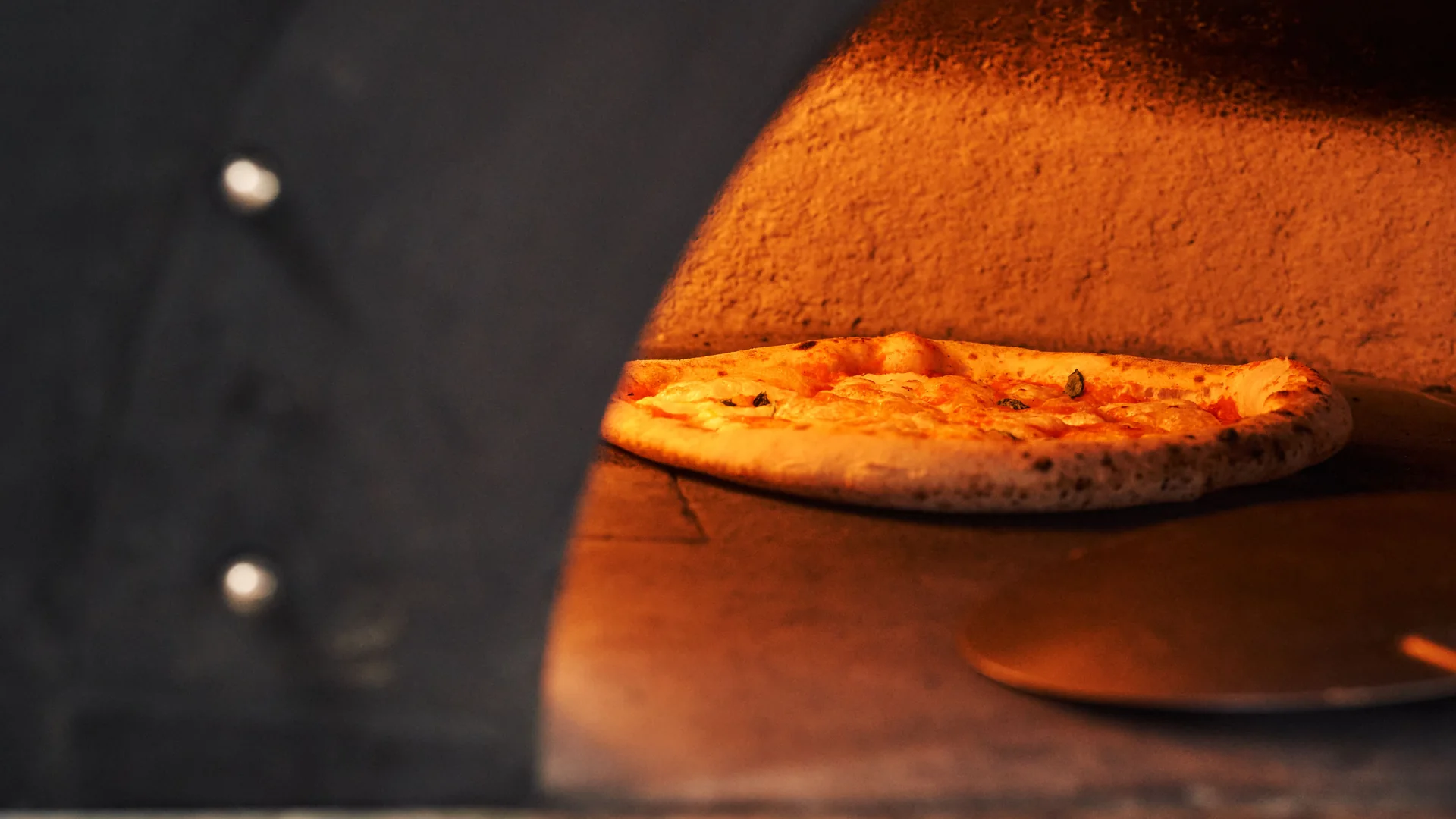 A pizza with a blistered, charred crust resting on the floor of a glowing wood-fired oven, a round metal peel lying beside it
